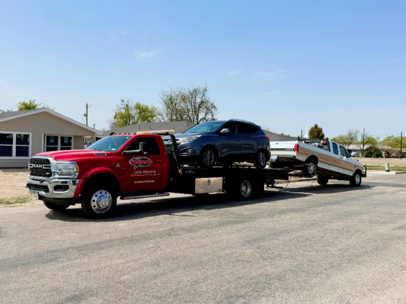 Red tow truck carrying a gray SUV and a white pickup truck on a sunny day.