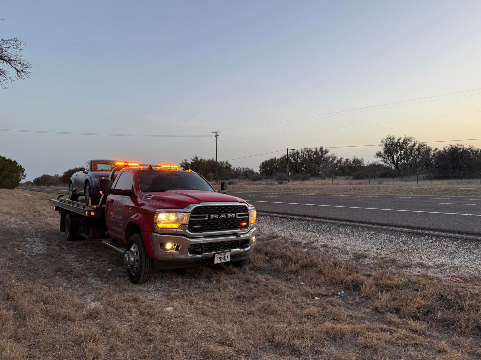 Red tow truck carrying a gray car on a roadside at dusk.