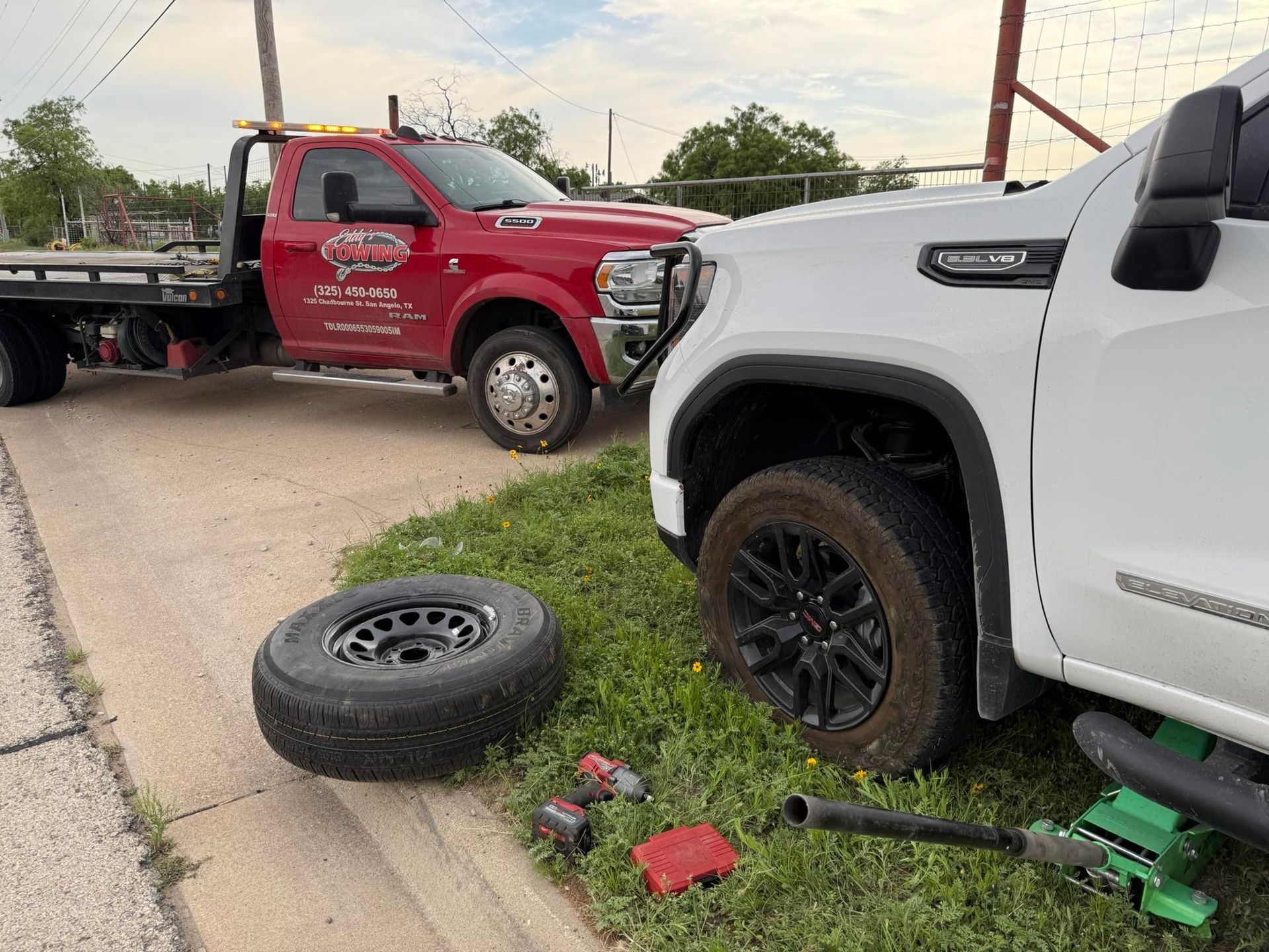 A white truck with a flat tire being assisted by a red tow truck on a roadside.