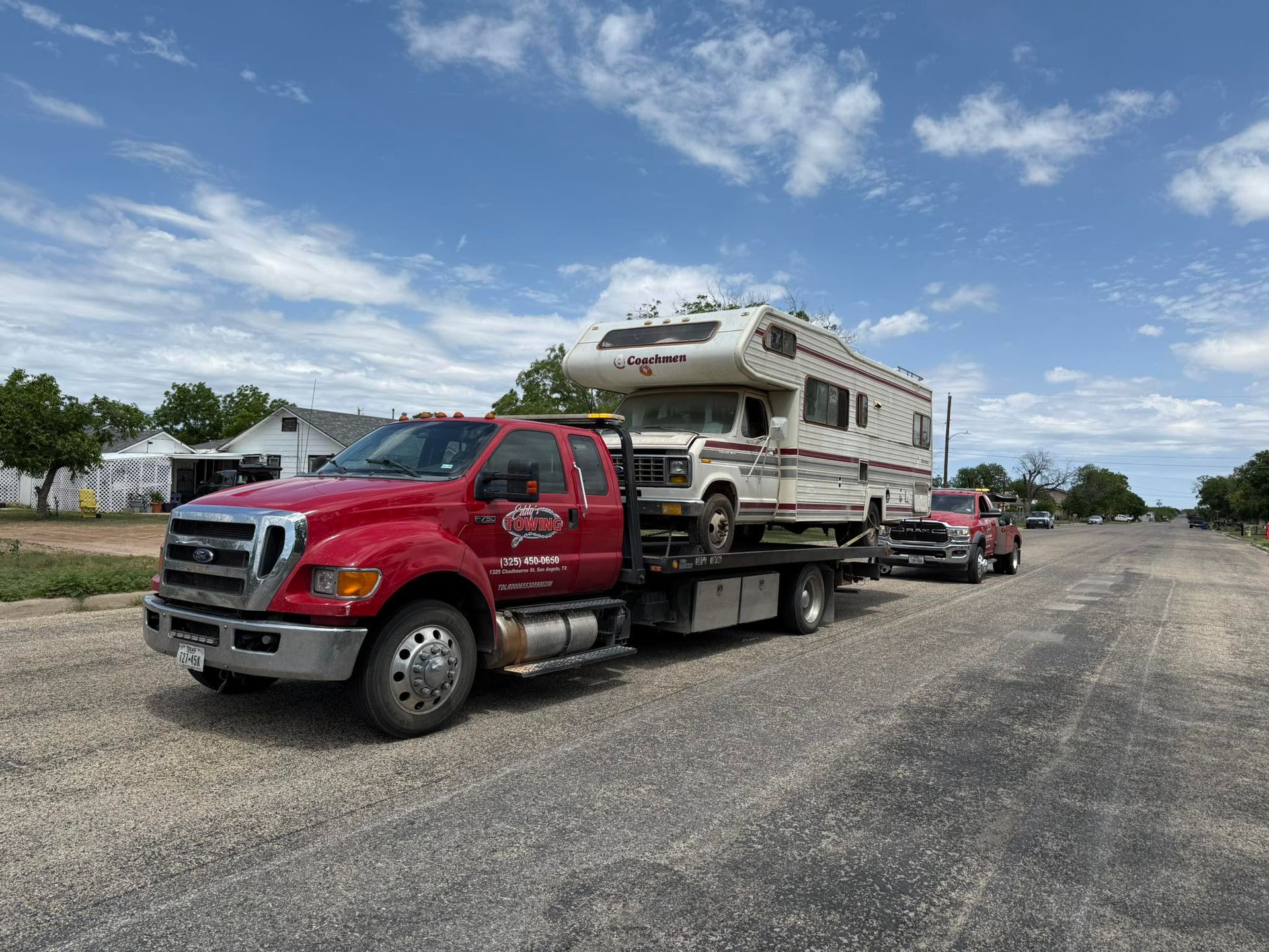 Red tow truck carrying a white and tan RV on a paved road under a cloudy blue sky.