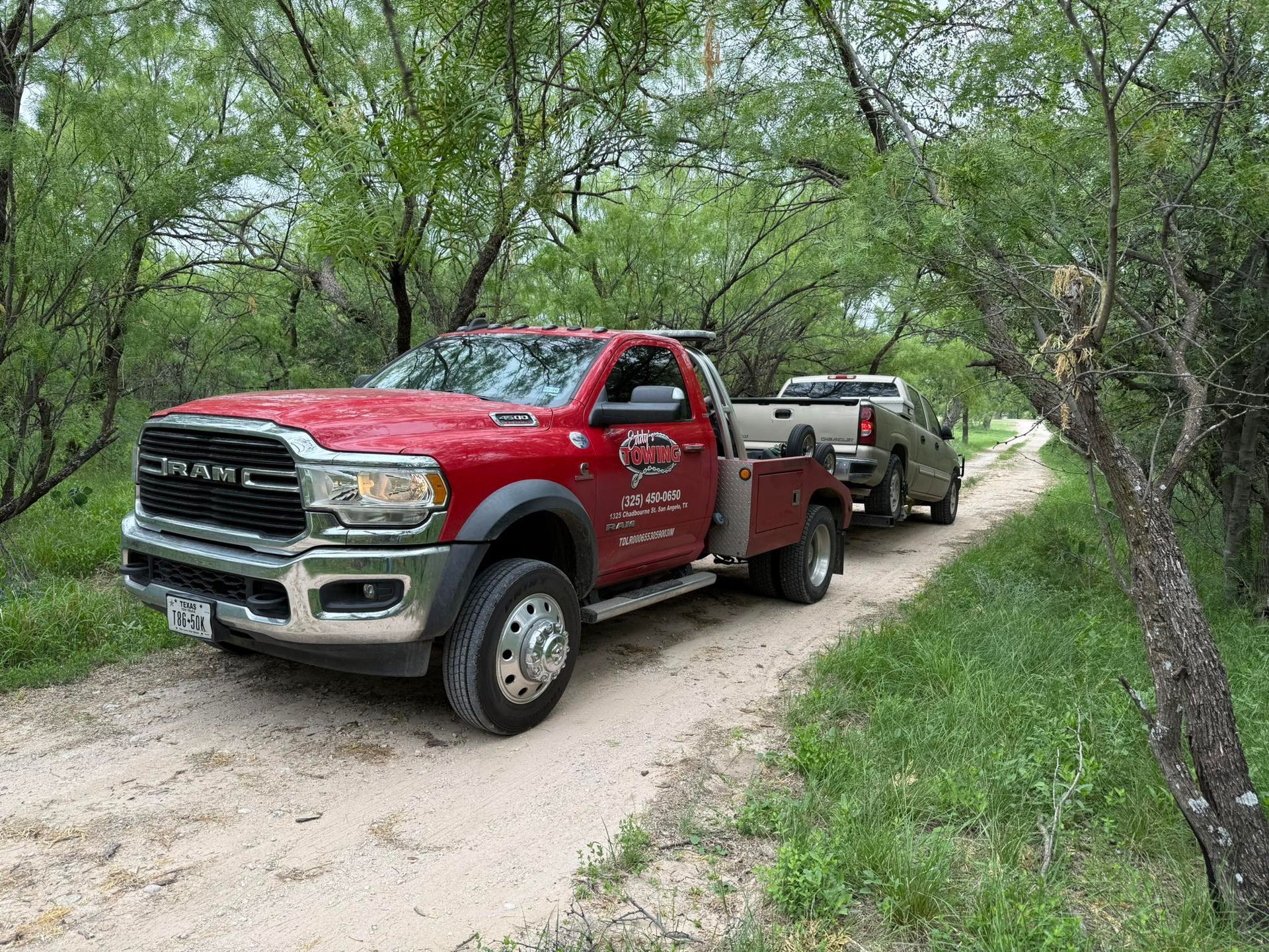 Red tow truck towing a smaller truck on a dirt road lined with trees.