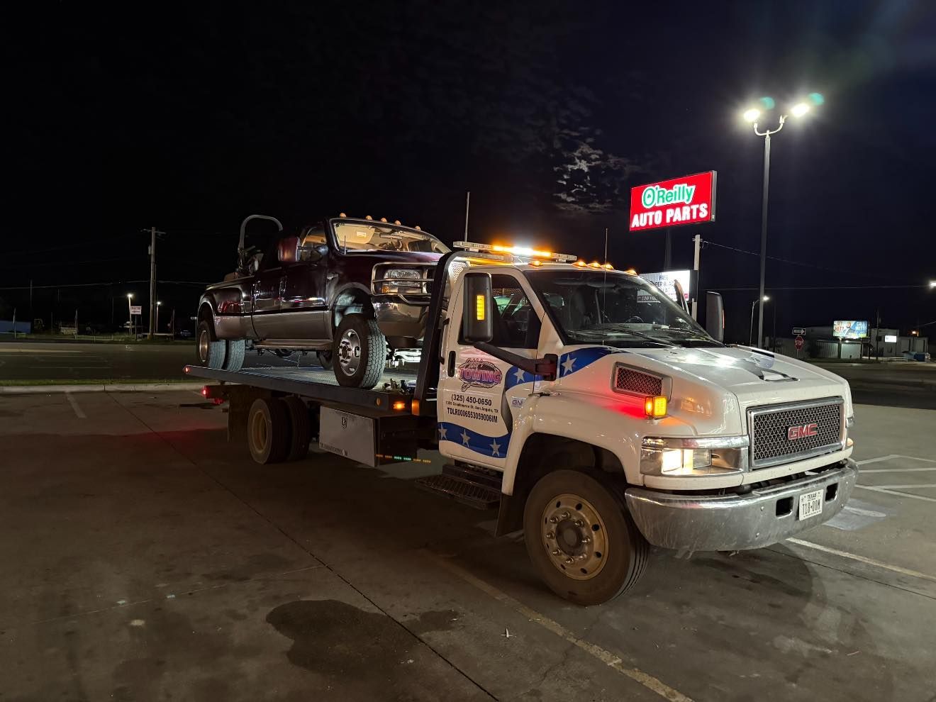 Tow truck with a car on its flatbed at night, parked near an auto parts store.