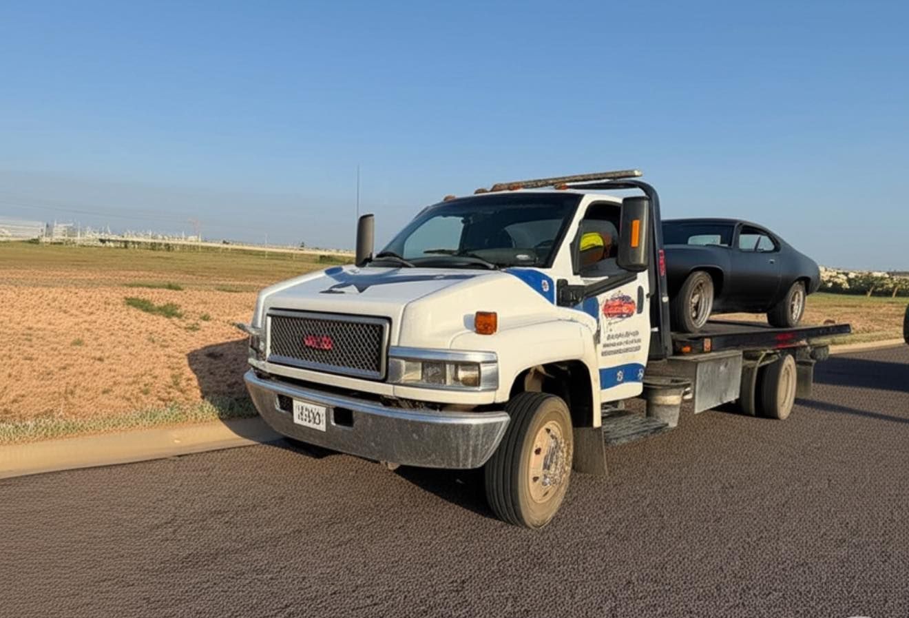 Tow truck hauling a classic black car on a sunny roadside. The truck is white with logos.
