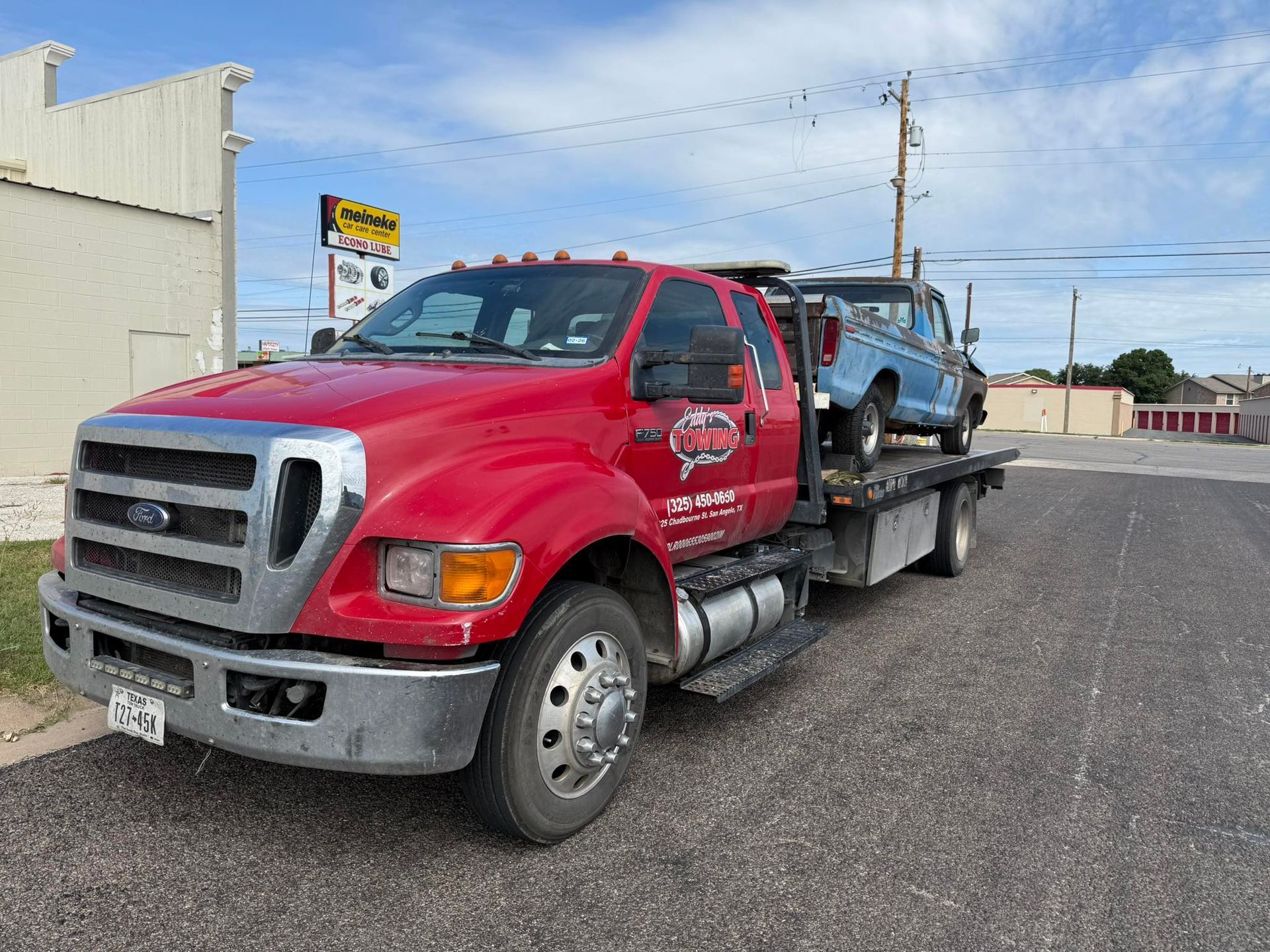 Red tow truck carrying a blue pickup truck on a city street under a partly cloudy sky.