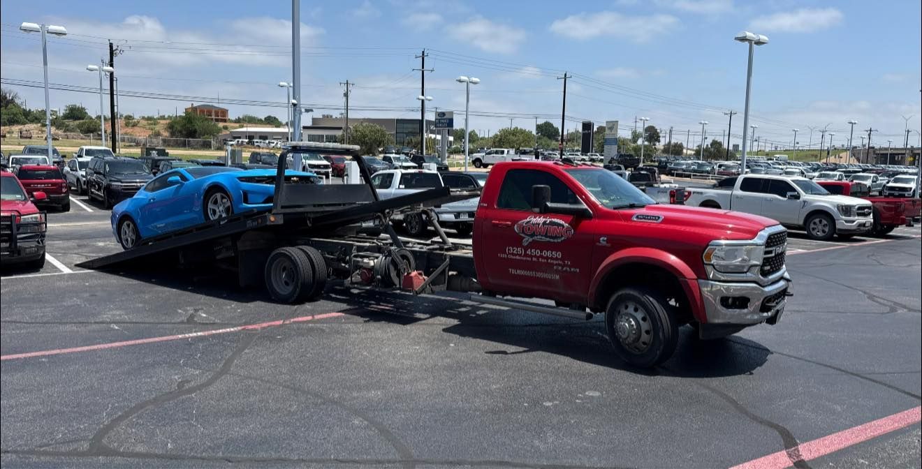 Red tow truck hauling a blue sports car in a parking lot on a sunny day.