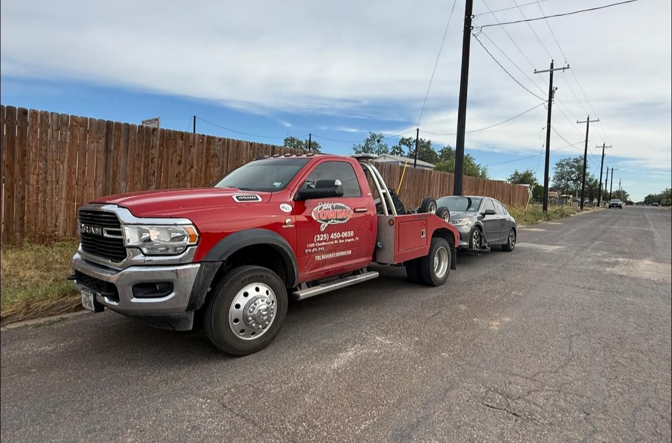 Red tow truck pulling a gray car on a street. Brown fence and power lines in the background.