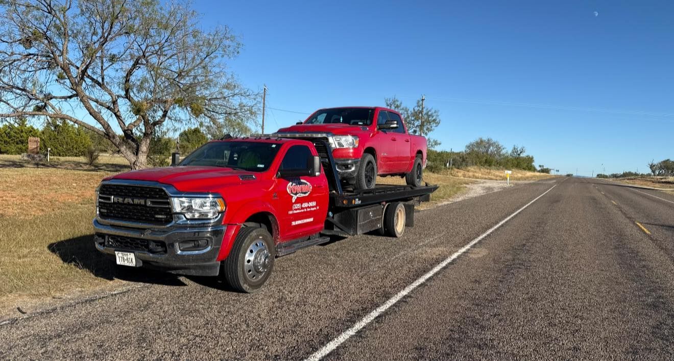 Red tow truck hauling a red pickup truck on a rural road under a blue sky.