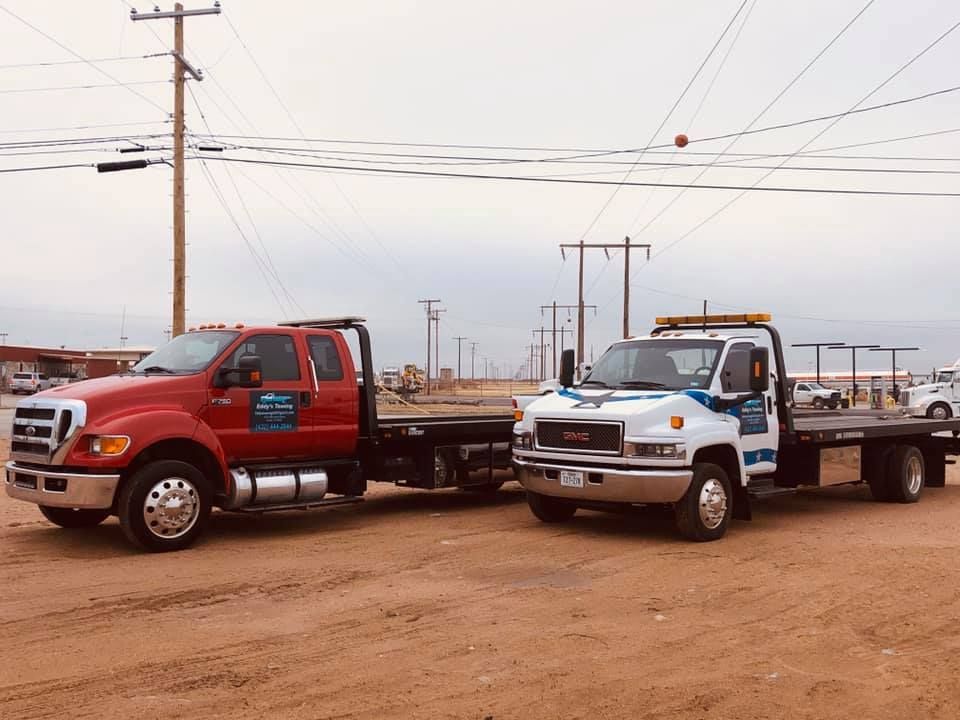 Two tow trucks parked on dirt road: red Ford, white GMC. Overhead power lines and cloudy sky.
