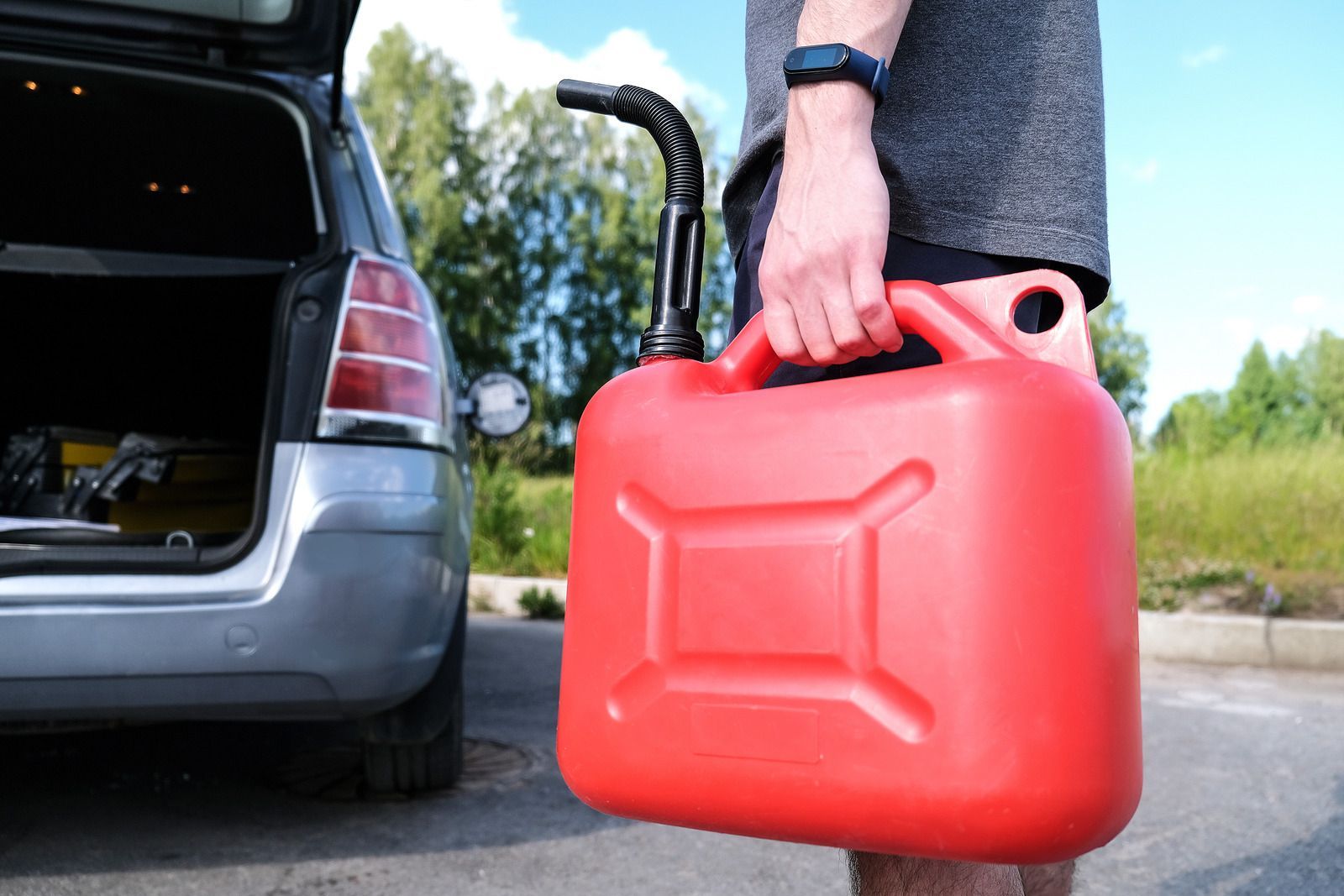 Person holding a red gas can near a car's open trunk on a paved road.