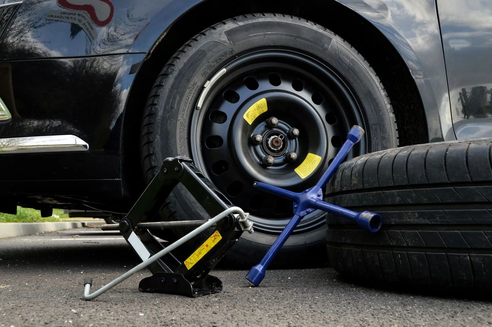 Car wheel with a blue and yellow tire jack beside a lifted black car on pavement