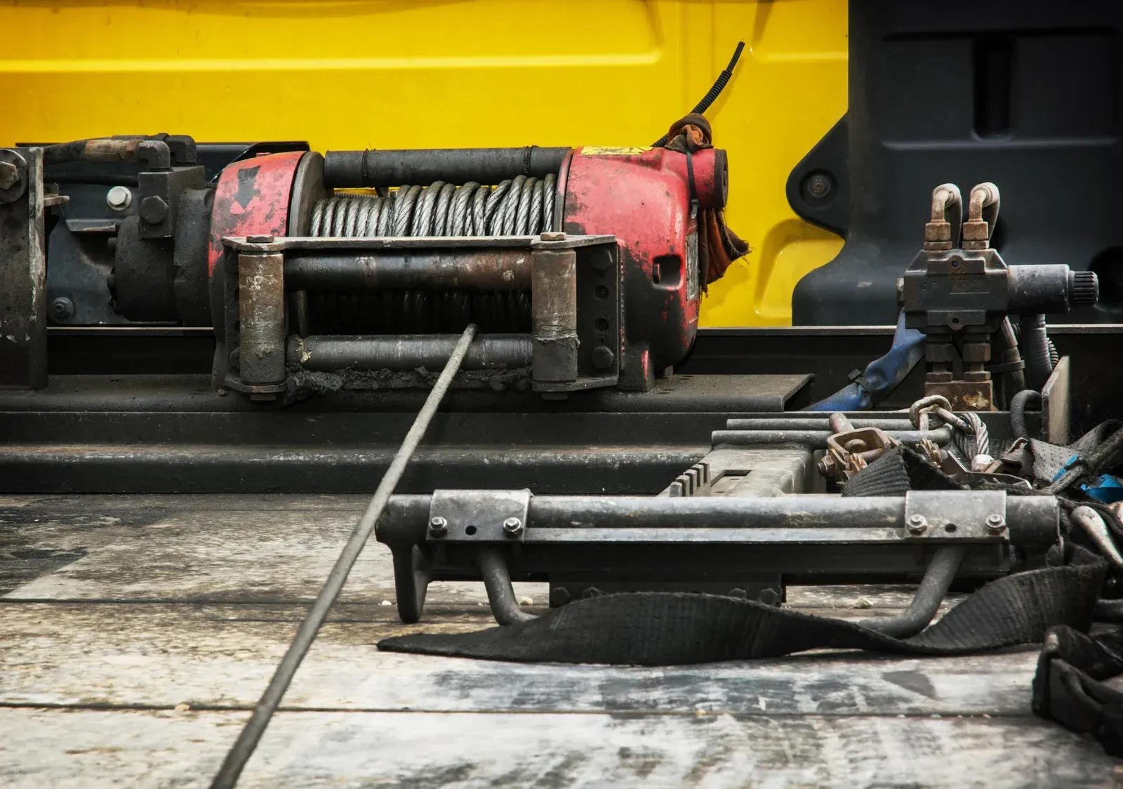 A weathered winch on a yellow truck bed. Red and black metal with coiled cable.