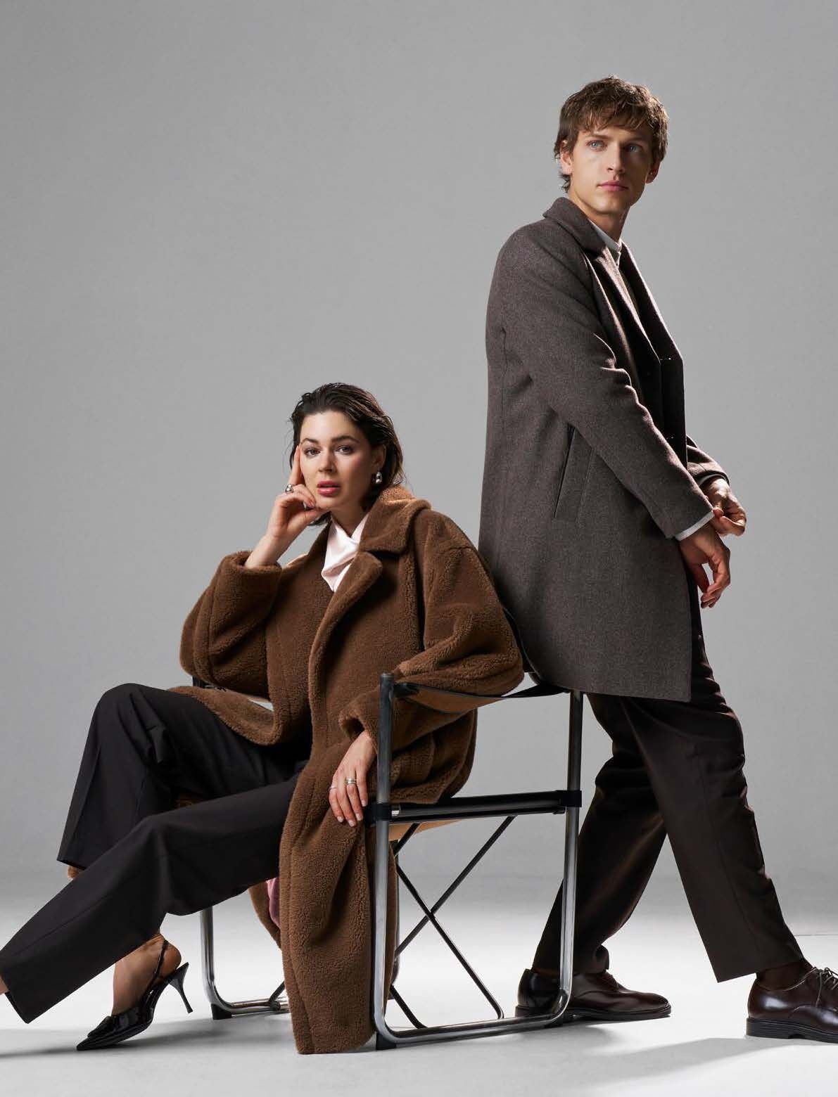 Frau sitzt, Mann steht; beide tragen Mäntel und posieren vor einem grauen Hintergrund.