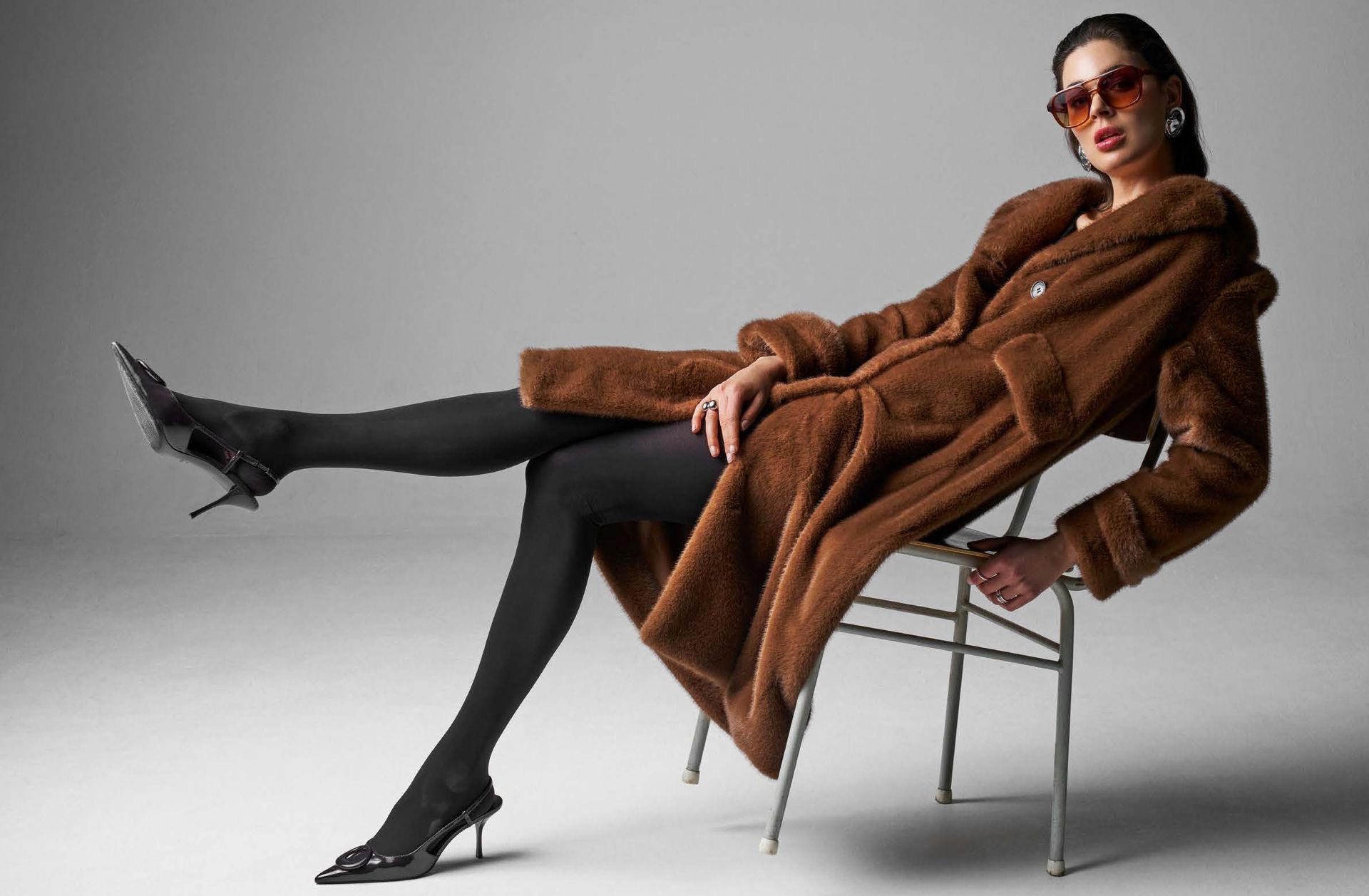 Frau in braunem Mantel, schwarzer Strumpfhose und High Heels, sitzt auf einem Metallstuhl, trägt eine Sonnenbrille.