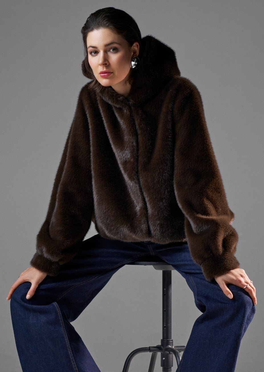 Eine Frau in einer braunen Pelzjacke mit Kapuze und blauen Jeans sitzt auf einem Hocker und schaut den Betrachter an.