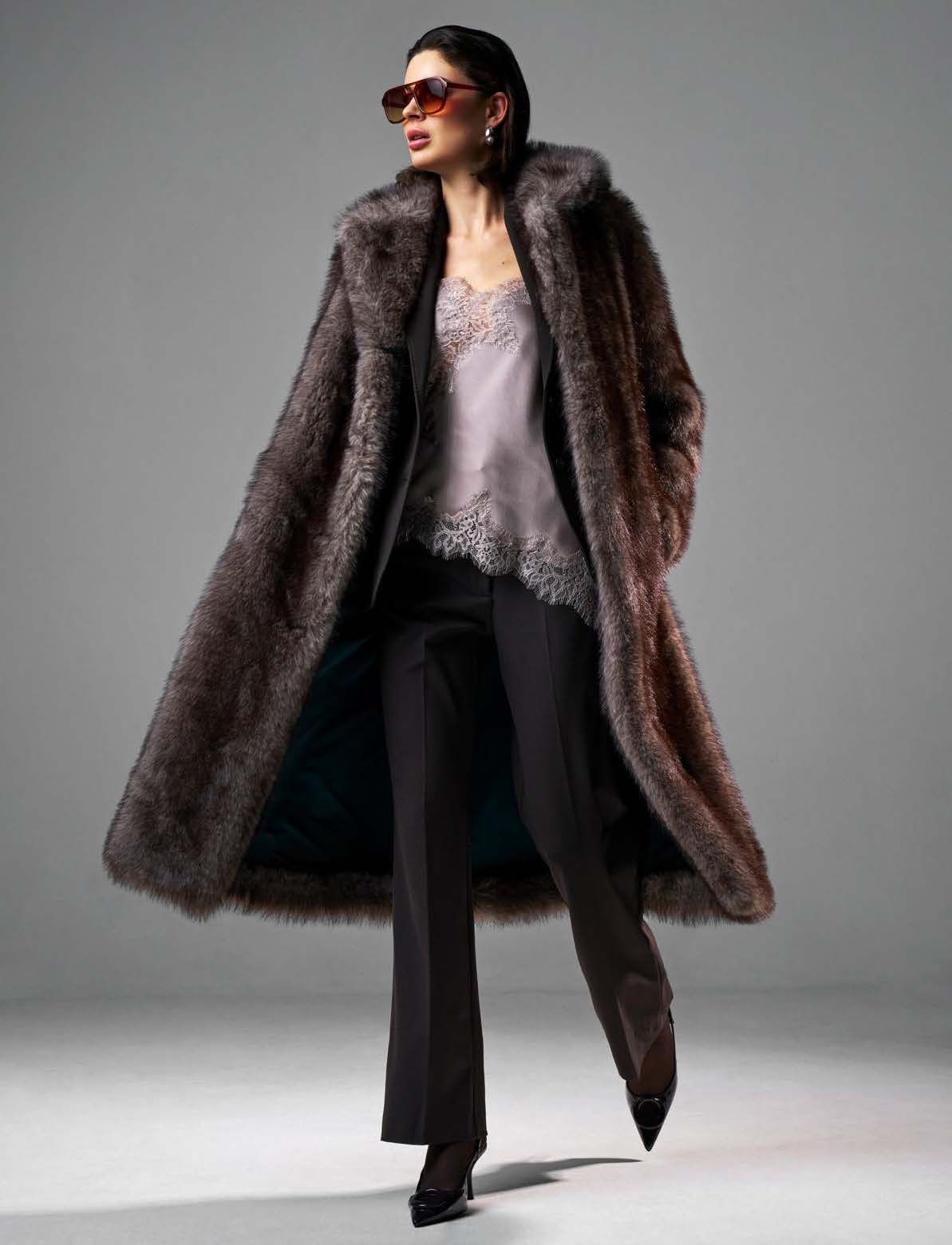 Frau in langem, braunem Pelzmantel, schwarzer Hose und High Heels, mit Sonnenbrille. Posiert vor grauem Hintergrund.