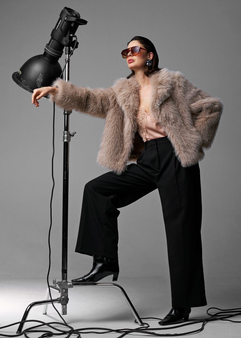 Eine Frau in flauschiger Jacke, Sonnenbrille und schwarzer Hose posiert im Studiolicht.