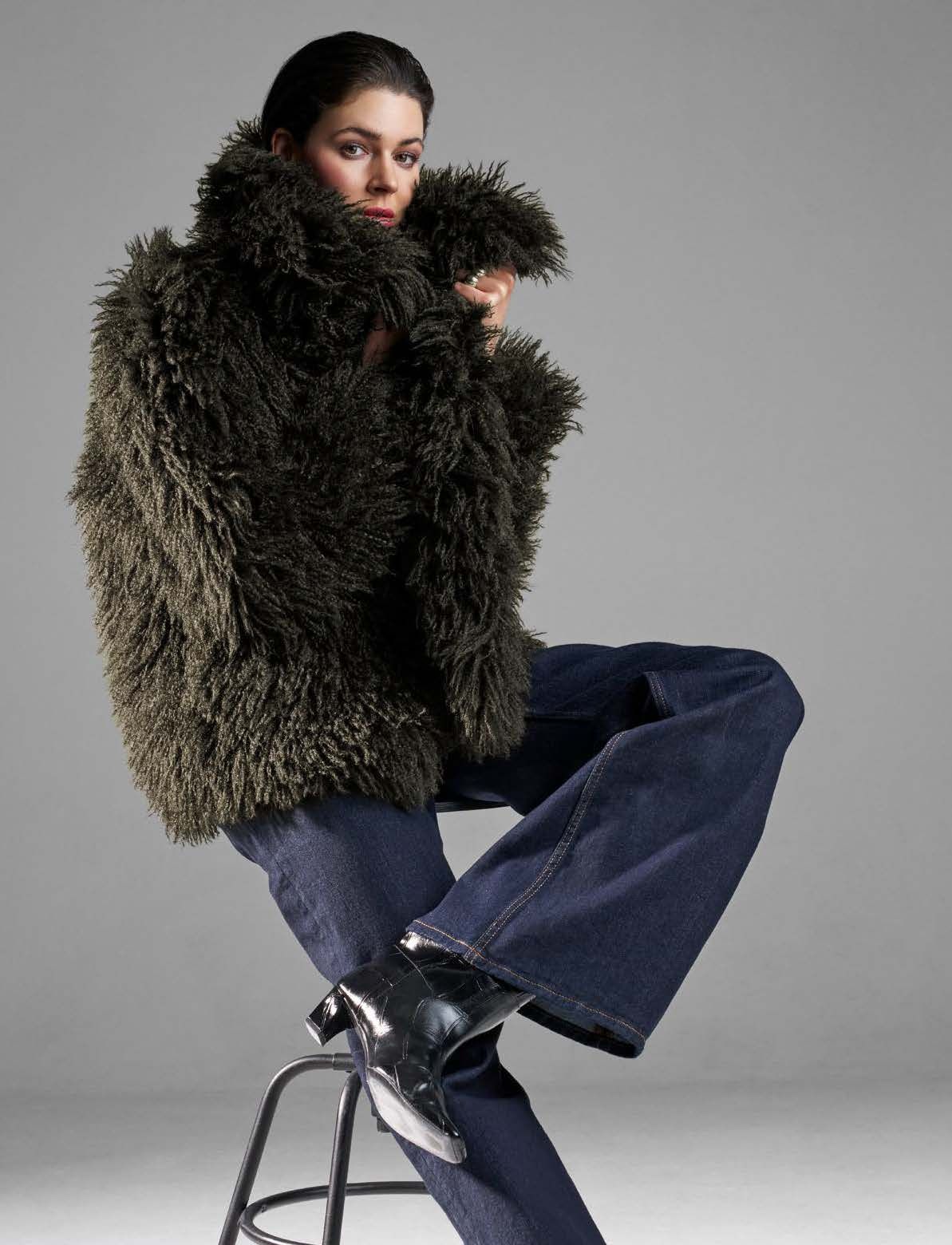 Frau in flauschiger olivgrüner Jacke, die auf einem Hocker sitzt, trägt blaue Jeans und schwarze Stiefel.