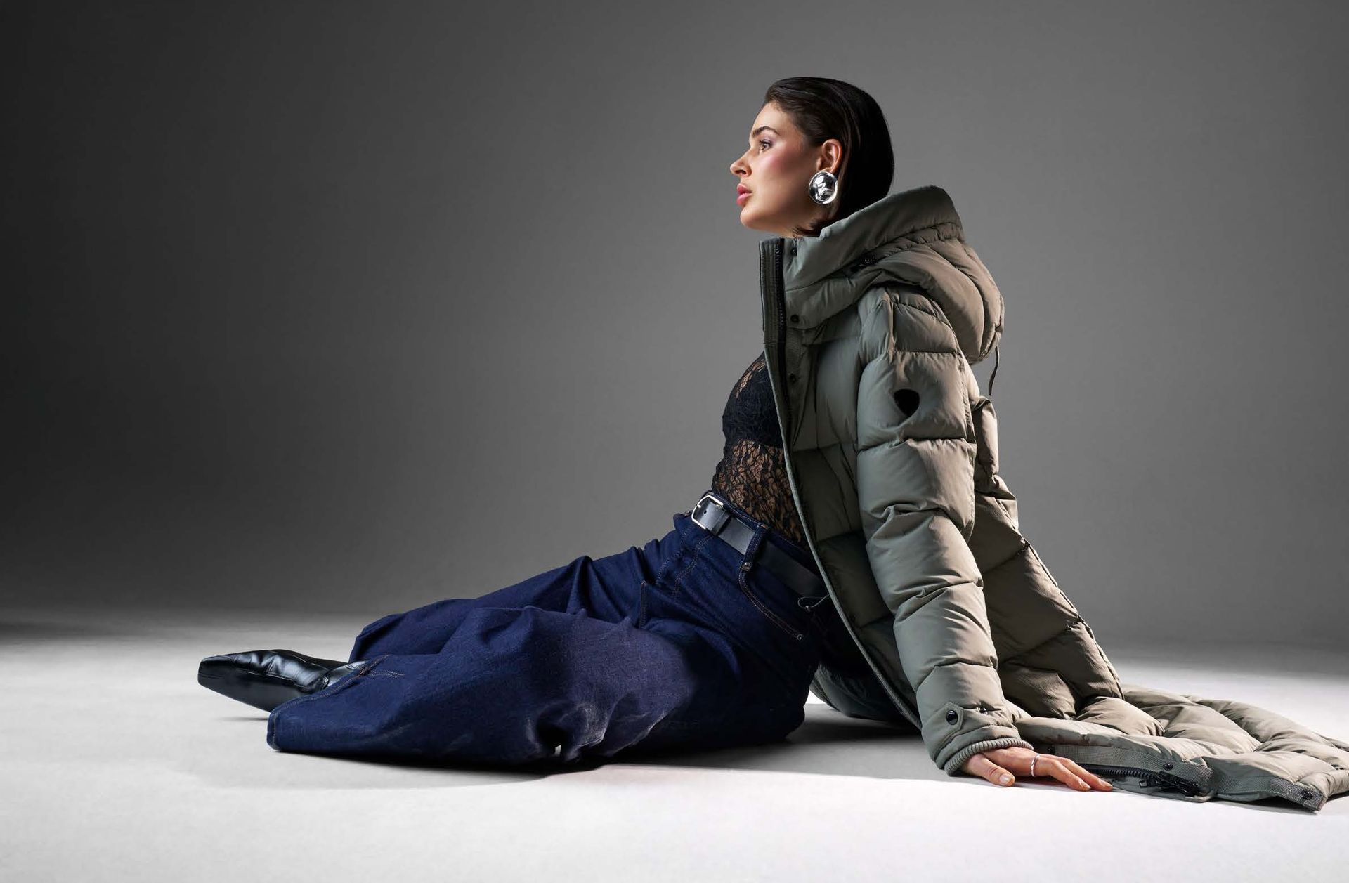 Frau in übergroßem Daunenmantel, Jeans und Ohrringen, sitzt auf einem grauen Boden, den Kopf nach hinten geneigt.