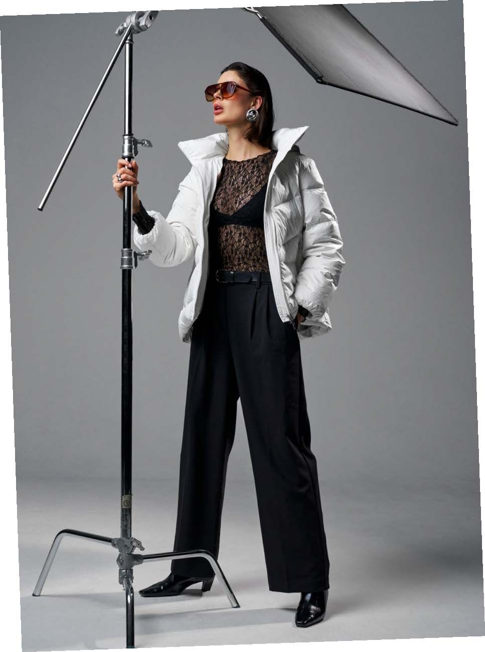 Eine Frau mit Sonnenbrille, weißer Jacke und schwarzer Hose posiert an einem Lampenstativ vor einem grauen Hintergrund.