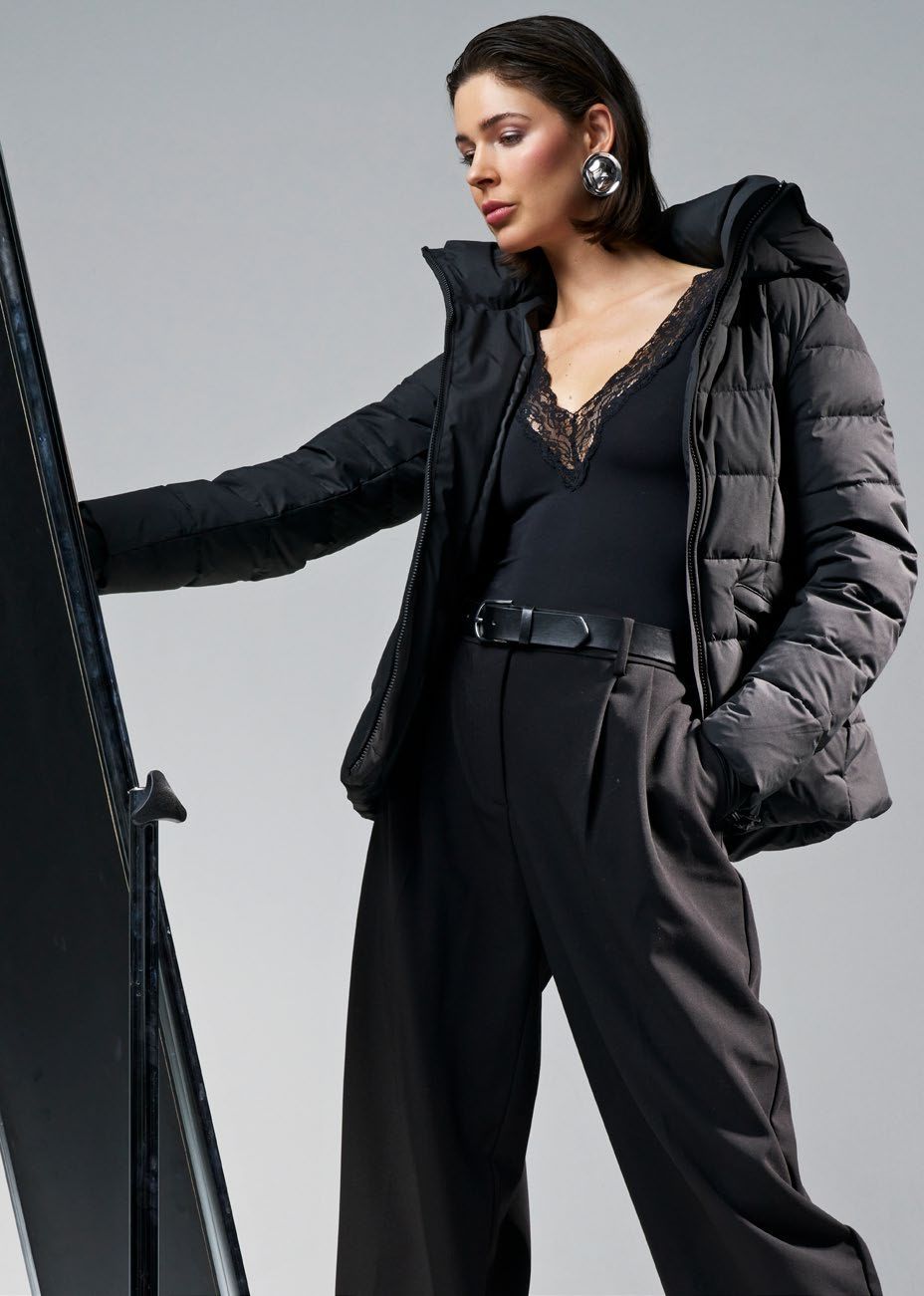 Frau in schwarzem Outfit, die eine Daunenjacke mit Kapuze trägt, posiert vor einem Spiegel.