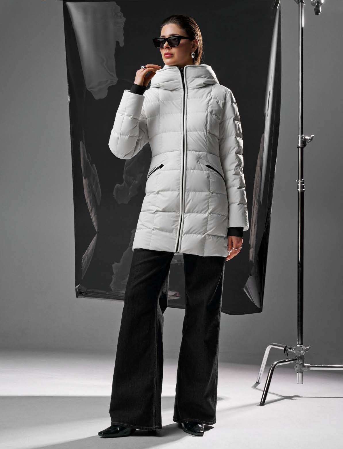 Frau in weißem Kapuzenmantel und schwarzer Hose posiert in einem Studio.