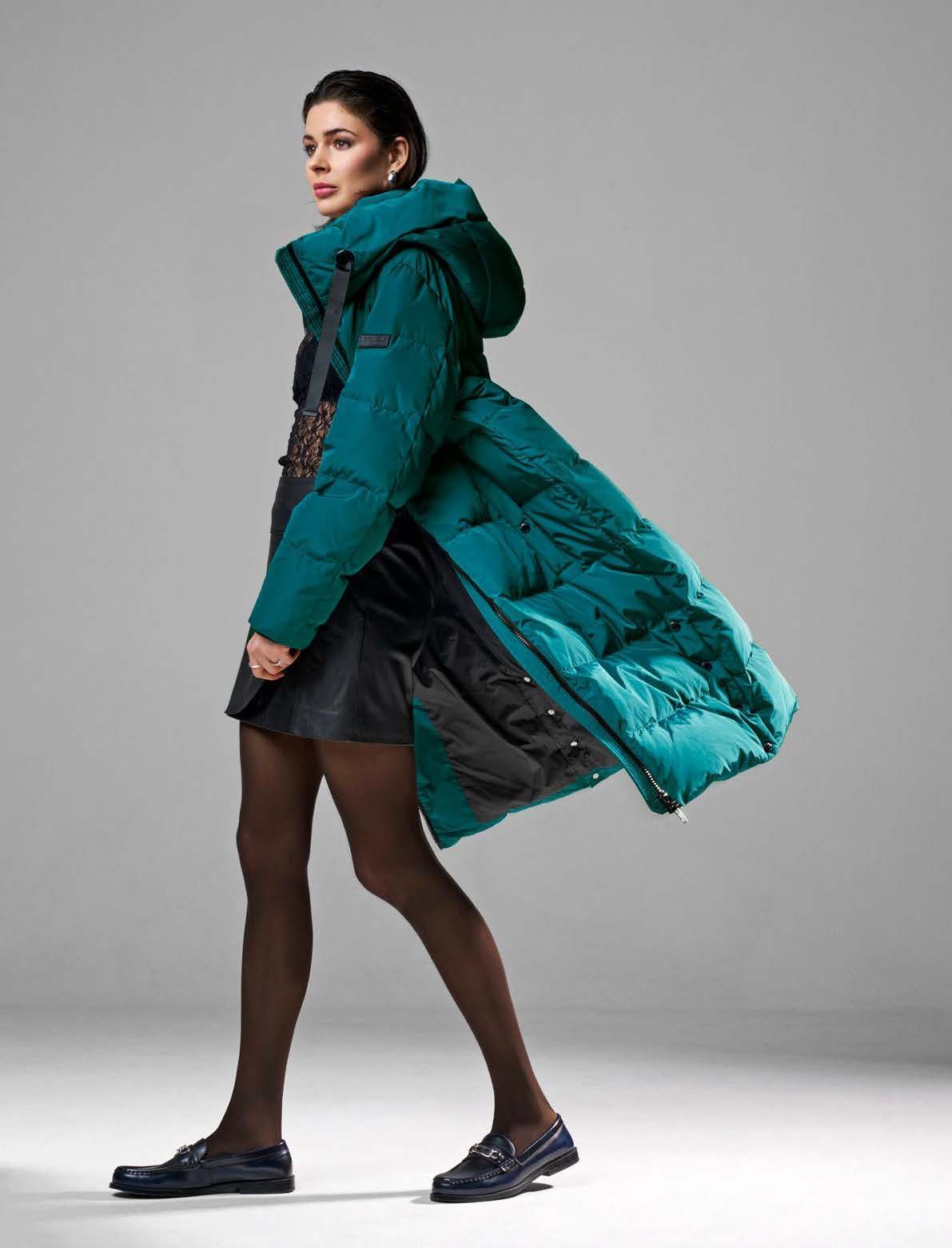 Eine Frau in einem langen, türkisfarbenen Daunenmantel, einem schwarzen Kleid und einer dunklen Strumpfhose geht.