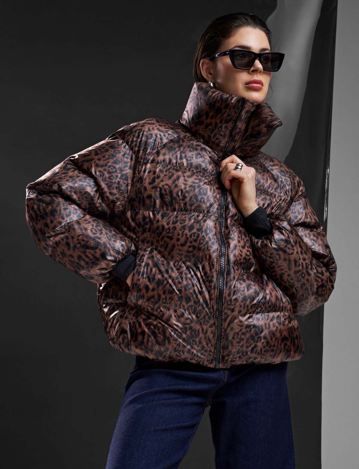 Frau in Leopardenmuster-Daunenjacke, Sonnenbrille und dunkler Hose. Steht vor einem dunklen Hintergrund.