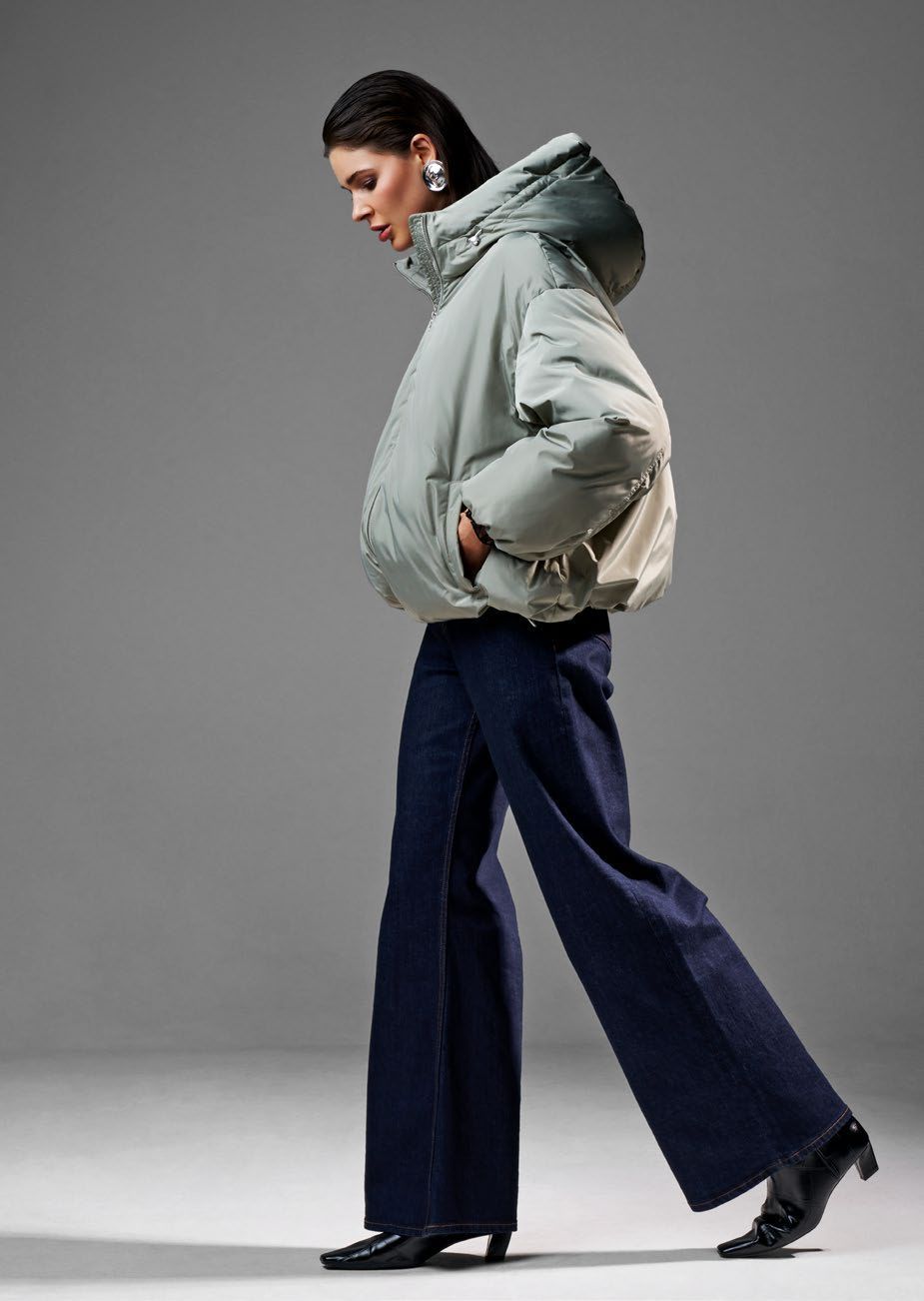 Frau in grüner Daunenjacke und weit geschnittener blauer Jeans geht, Hände in den Taschen, Studio-Kulisse.