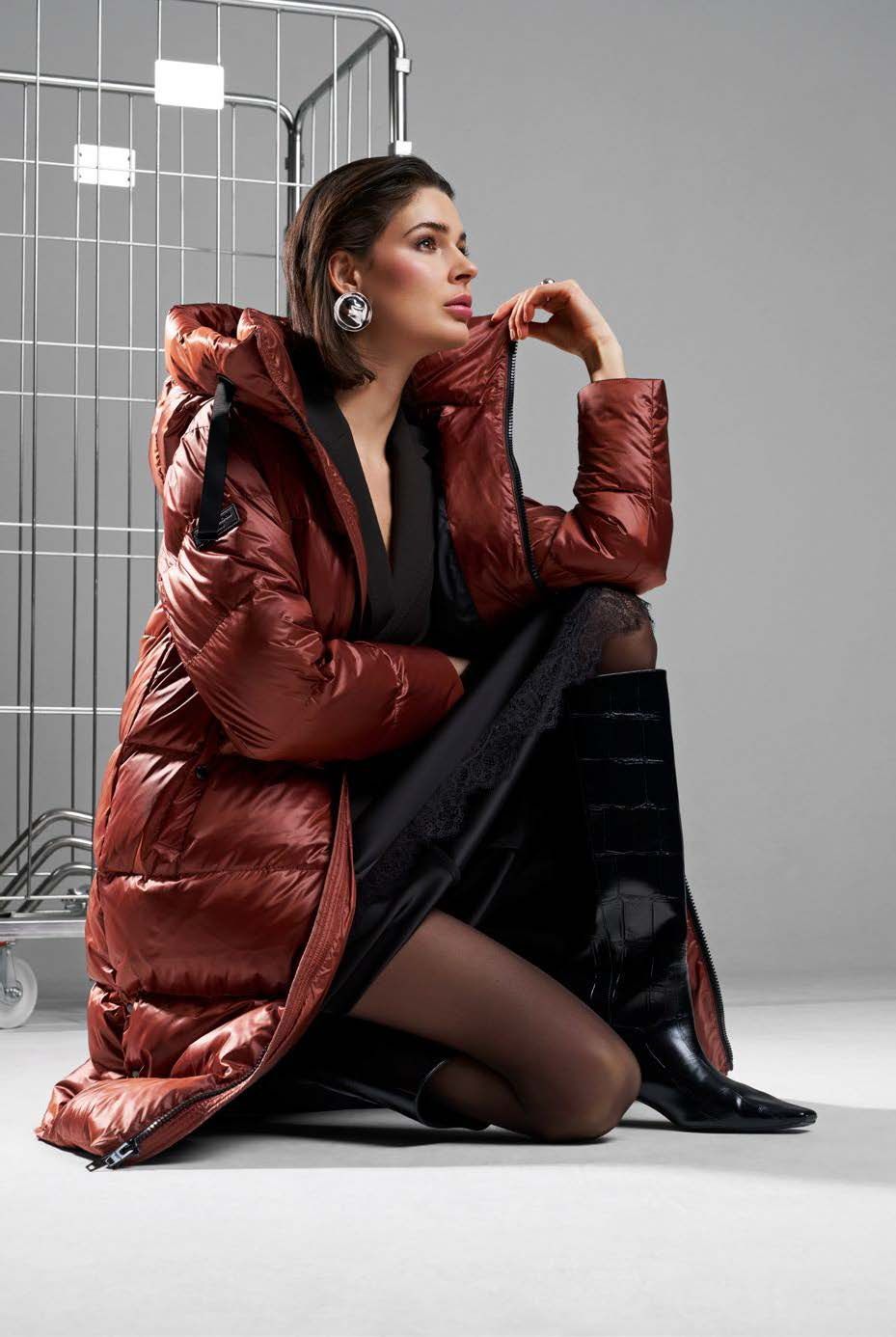 Eine Frau in einem langen, roten Daunenmantel kniet und blickt zur Seite. Hinter ihr steht ein Metallgestell.