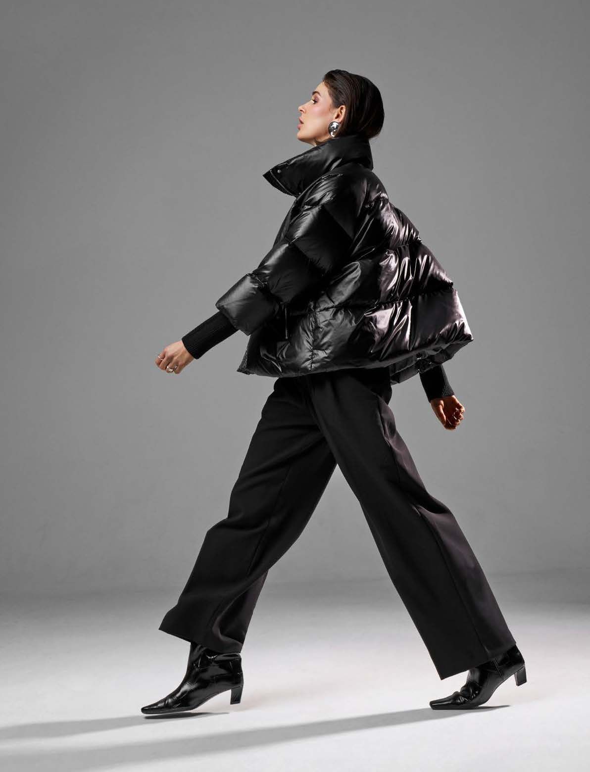 Frau in schwarzer Daunenjacke, Hose und Stiefeln geht. Grauer Hintergrund.