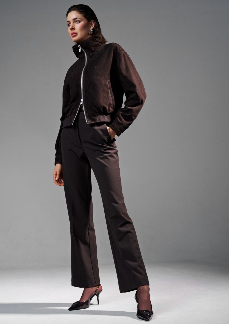 Eine Frau in einem braunen Hosenanzug und High Heels posiert vor einem grauen Hintergrund.