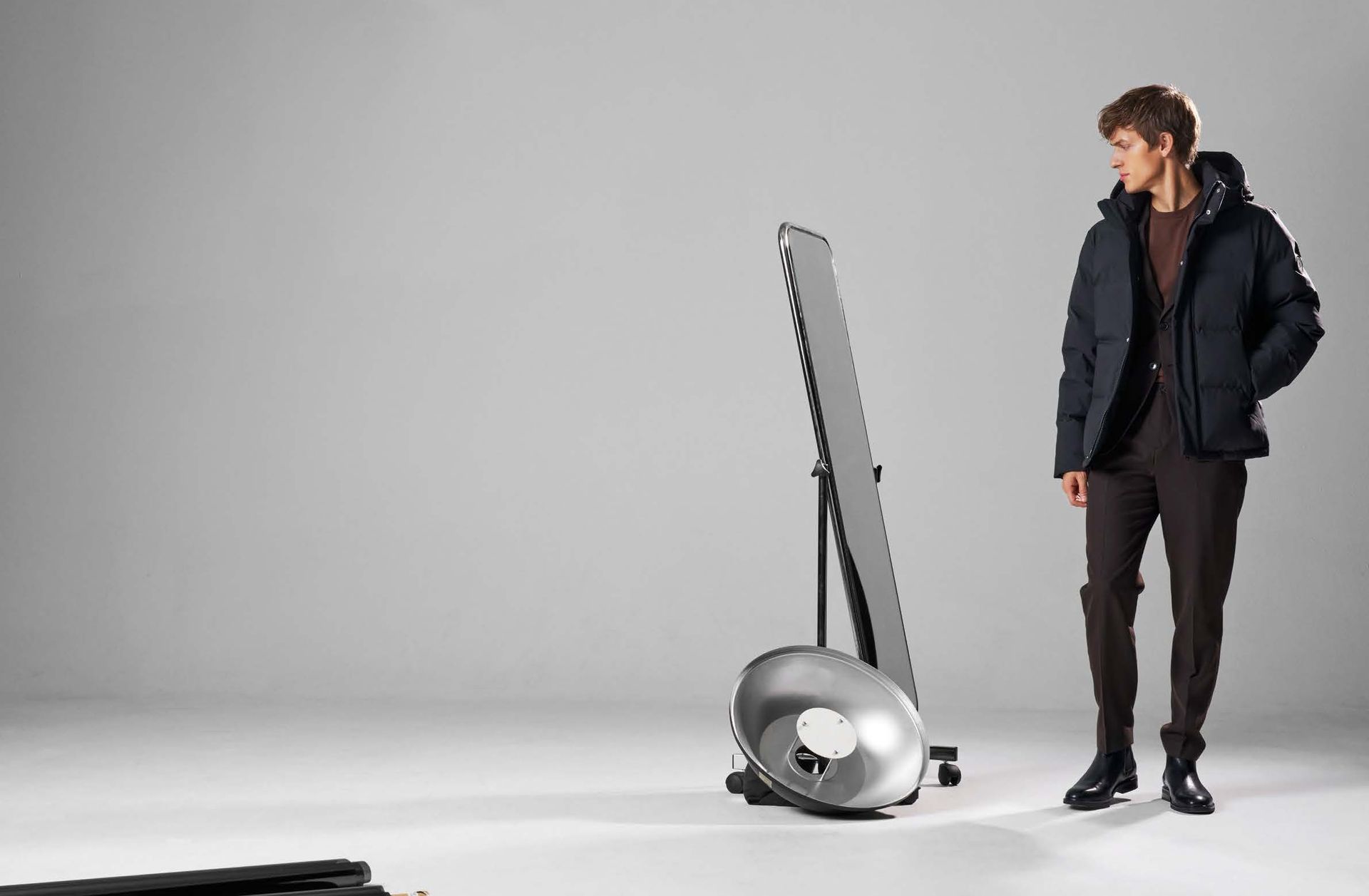 Ein Mann in blauer Jacke und brauner Hose blickt in einem Studio auf einen großen Spiegel und einen Reflektor.