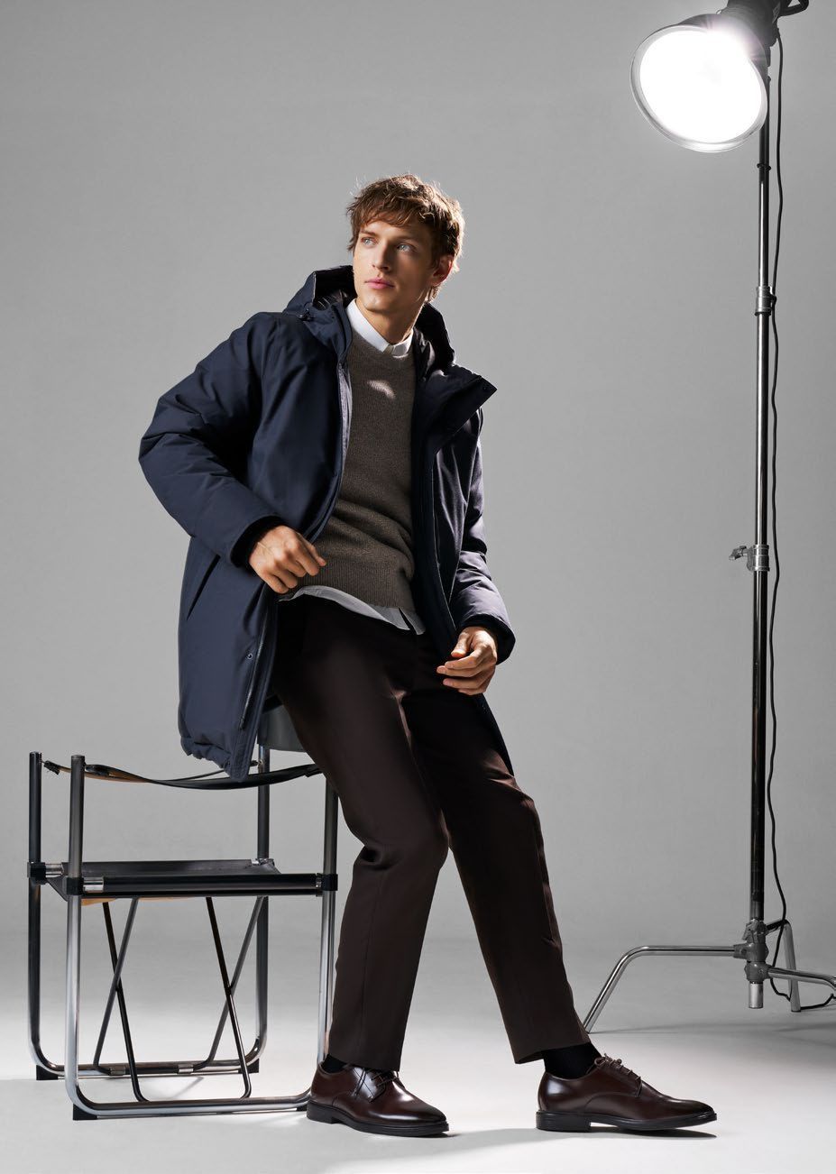 Ein Mann in einem dunkelblauen Mantel und braunen Hosen sitzt unter Studiobeleuchtung auf einem Stuhl.