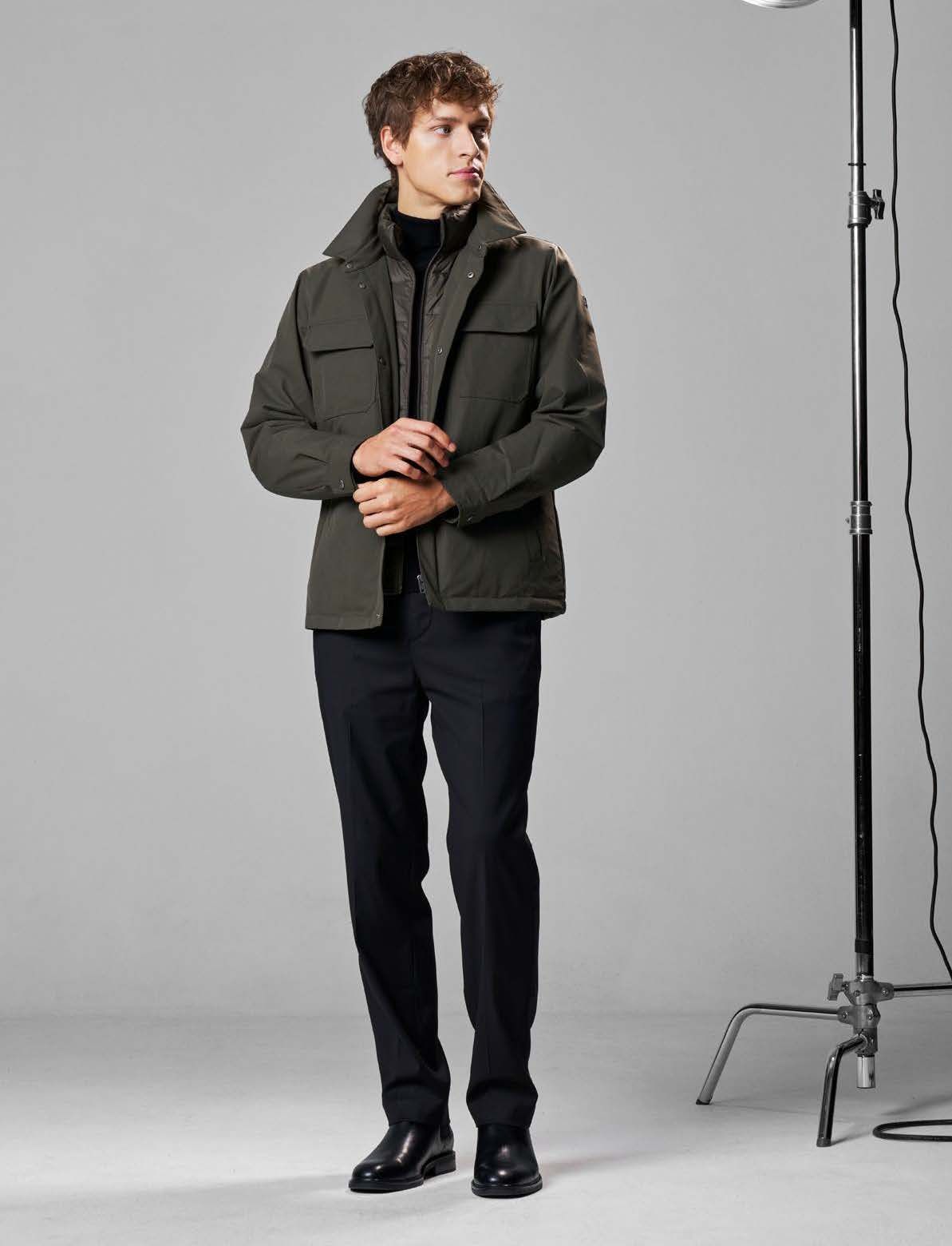 Mann in olivgrüner Jacke und schwarzer Hose, die Hände verschränkt, in einem Studio.