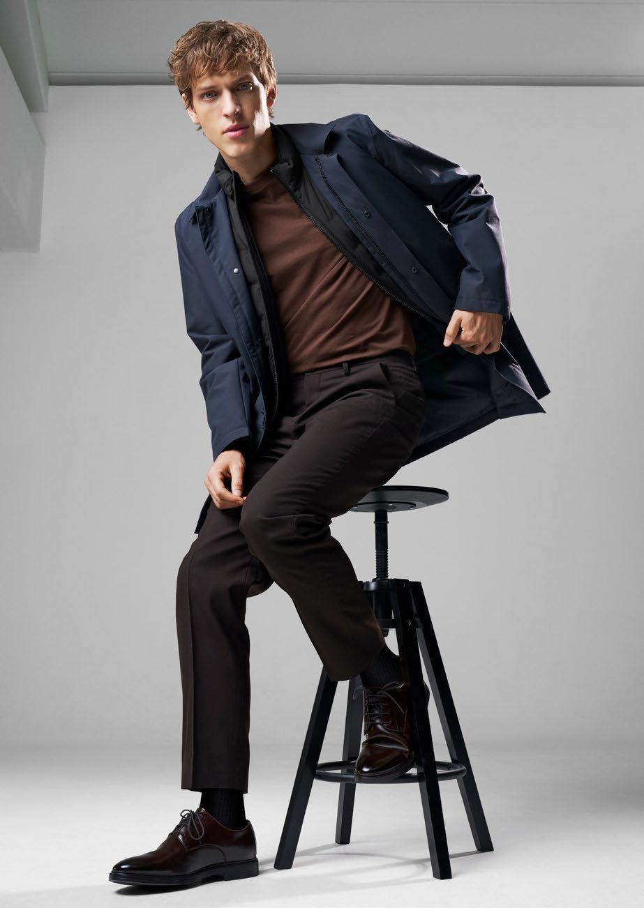 Ein Mann sitzt auf einem Hocker; er trägt einen blauen Mantel, ein braunes Hemd und eine dunkle Hose.