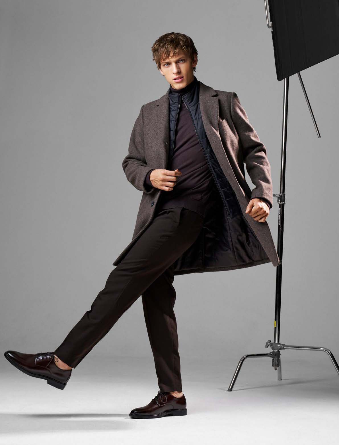Mann in Mantel und Hose posiert in einem Studio. Er trägt einen langen Mantel, eine dunkle Hose und Anzugschuhe.