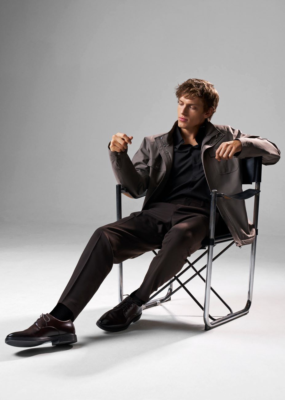 Mann im braunen Anzug, sitzend auf einem Regiestuhl, gestikulierend. Grauer Hintergrund.