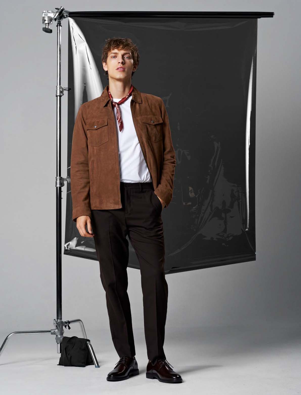 Mann in brauner Jacke, weißem Hemd, dunkler Hose und Schal posiert vor einer Studio-Kulisse.