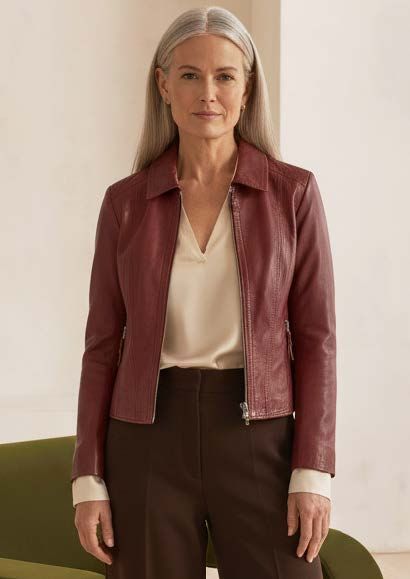 Frau in bordeauxroter Lederjacke, cremefarbenem Oberteil und brauner Hose, steht in einem Zimmer.