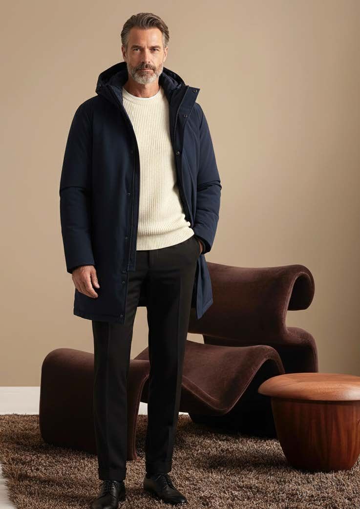 Mann in marineblauem Mantel, elfenbeinfarbenem Pullover und schwarzer Hose steht in einem Zimmer mit modernen Möbeln.