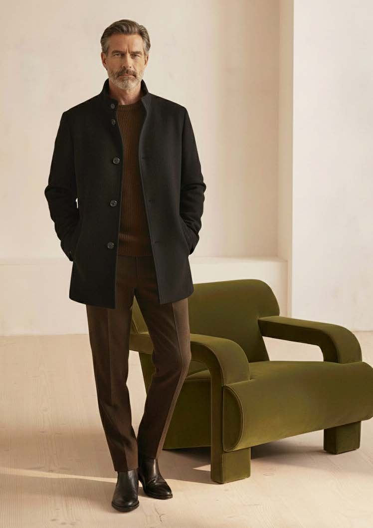 Mann in schwarzem Mantel, braunem Pullover, Hose und Stiefeln, der neben einem grünen Sessel steht.