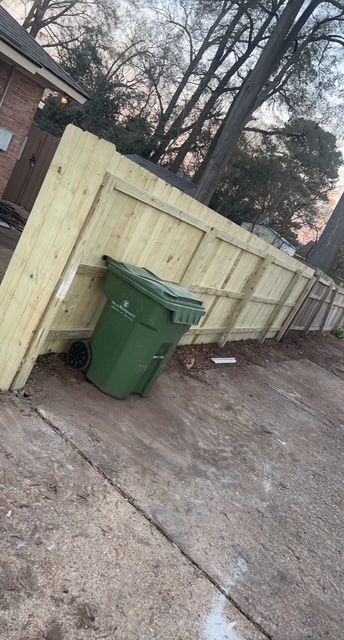 A green trash can is sitting next to a wooden fence.