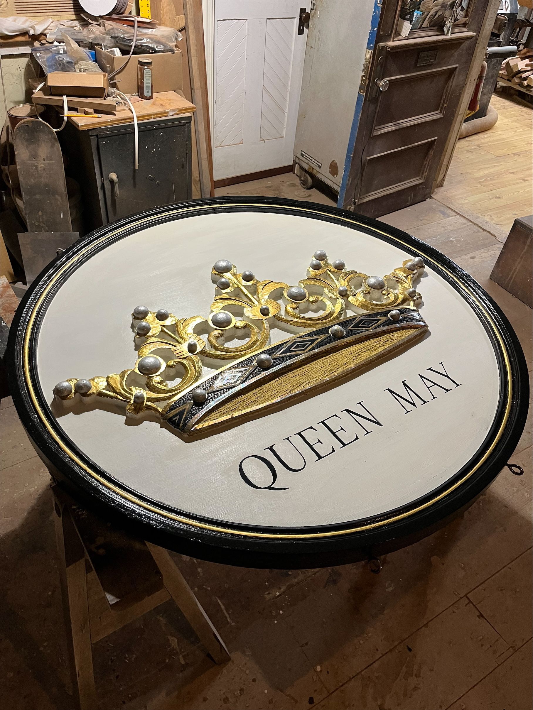 A round sign with a golden crown and Queen May text on a white background, black border designed by cape island woodworks in cape may, new jersey