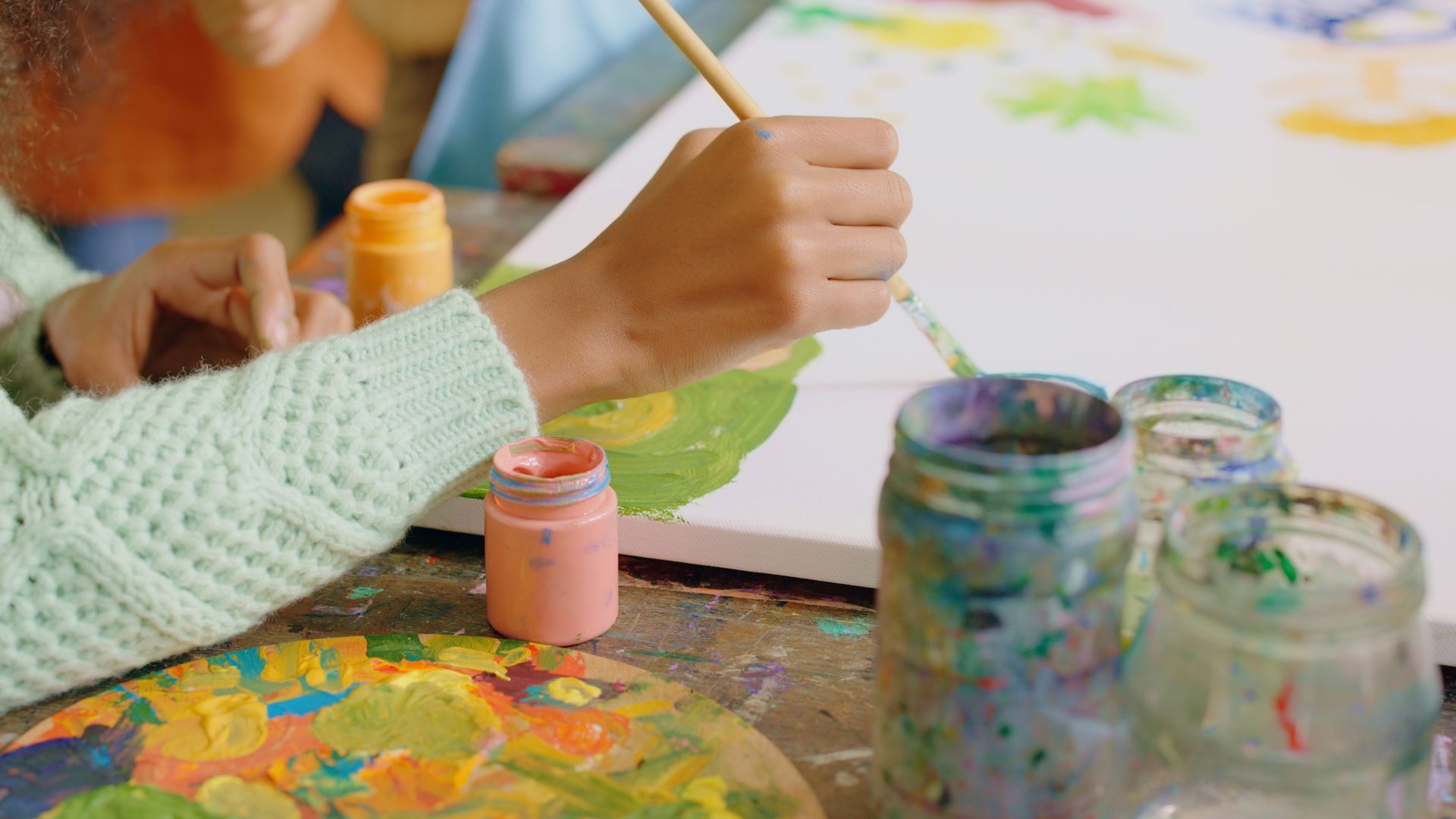 Person painting on a canvas with a brush, surrounded by paint jars and a palette.