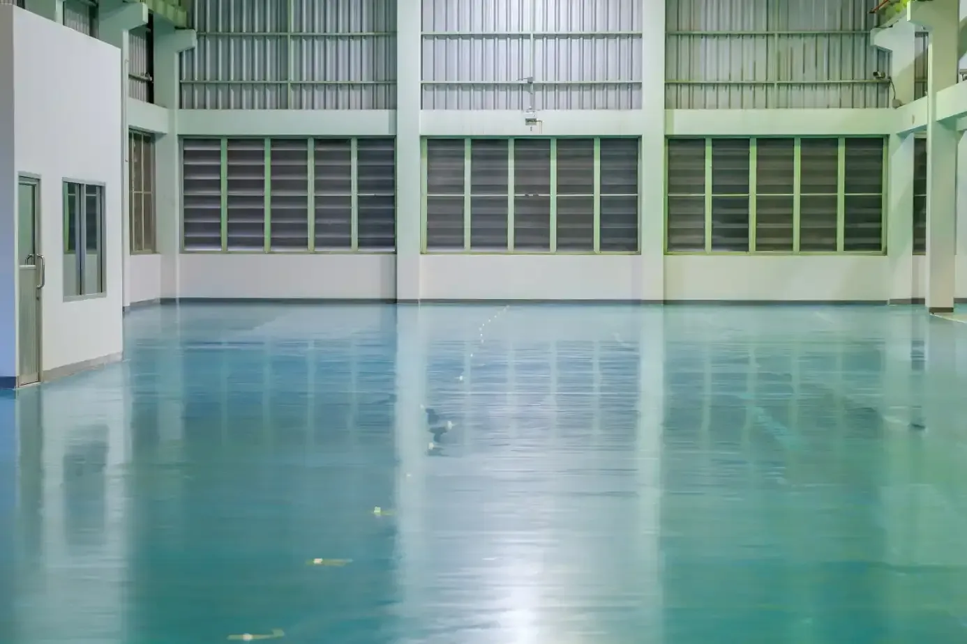 Empty industrial warehouse with teal-colored, reflective floor. Metal ceiling and wall windows.