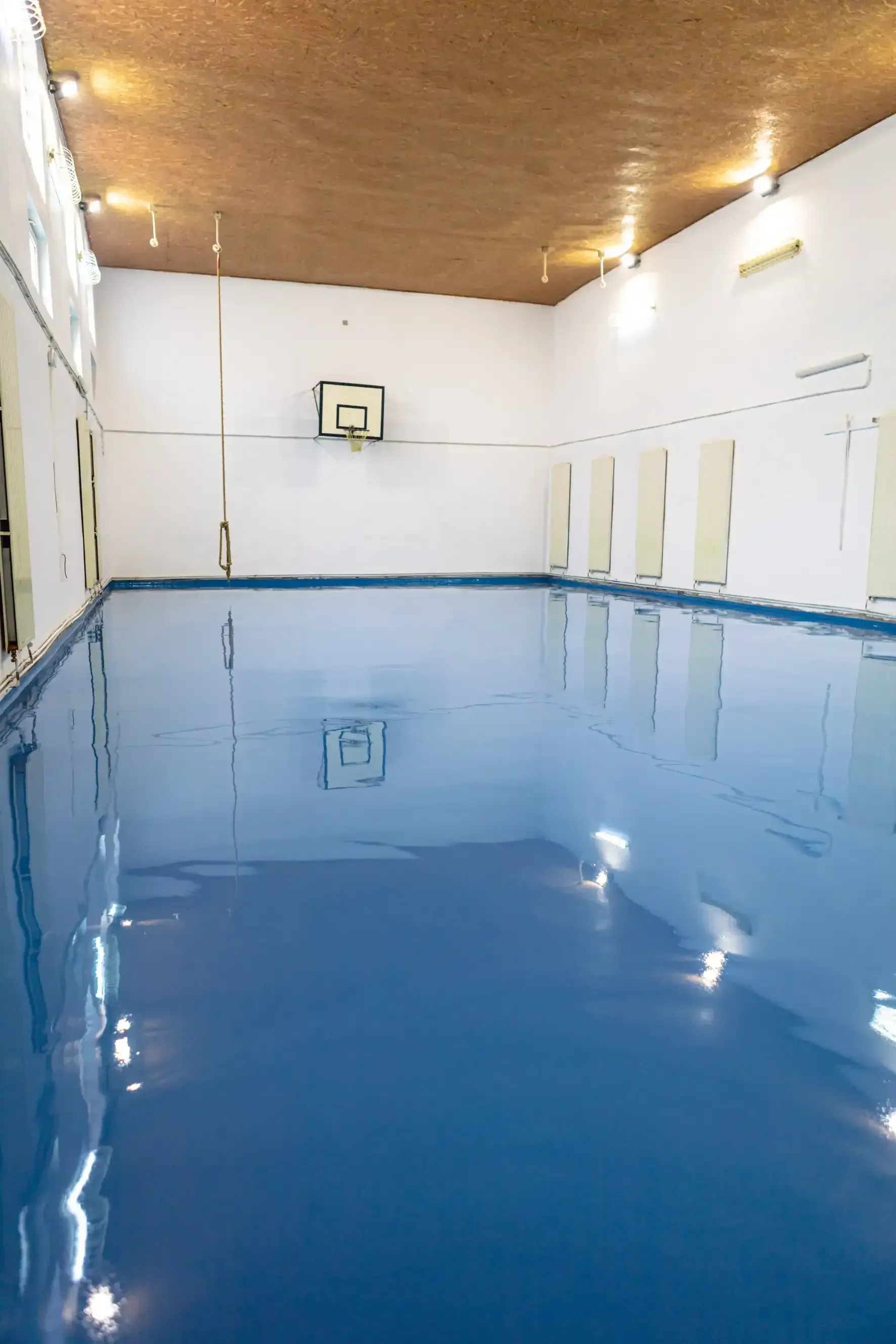 Indoor blue swimming pool with a basketball hoop on one end, white walls, and a light-brown ceiling.