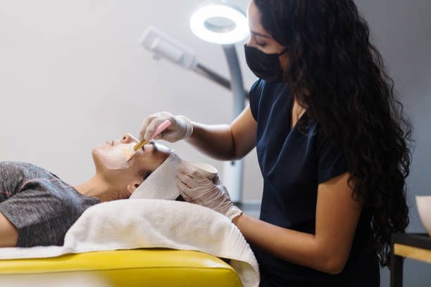 Esthetician wearing a mask and gloves applying facial mask to a client in a spa setting.