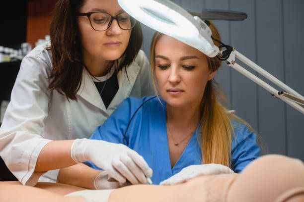 Two women, one in scrubs, one in a lab coat, focus on a medical procedure under a magnifying lamp.