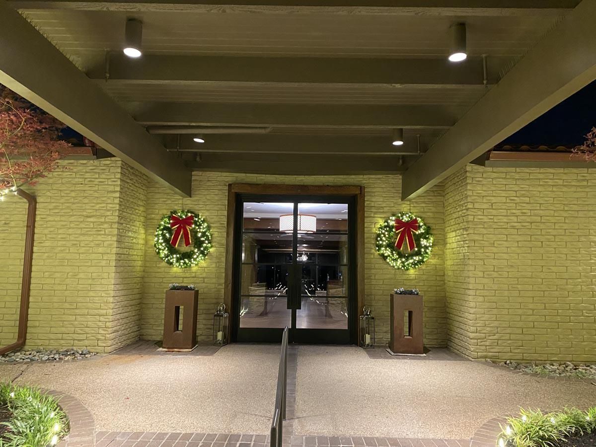 Entrance with wreaths and lights, brick walls, and glass doors at dusk.
