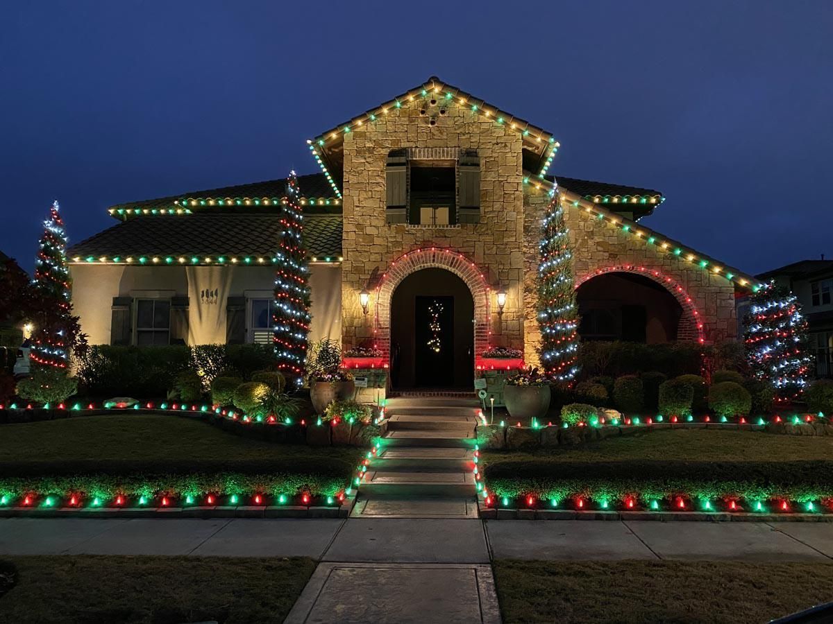 House with Christmas lights; green, red, and white lights adorn roof, bushes, and trees.